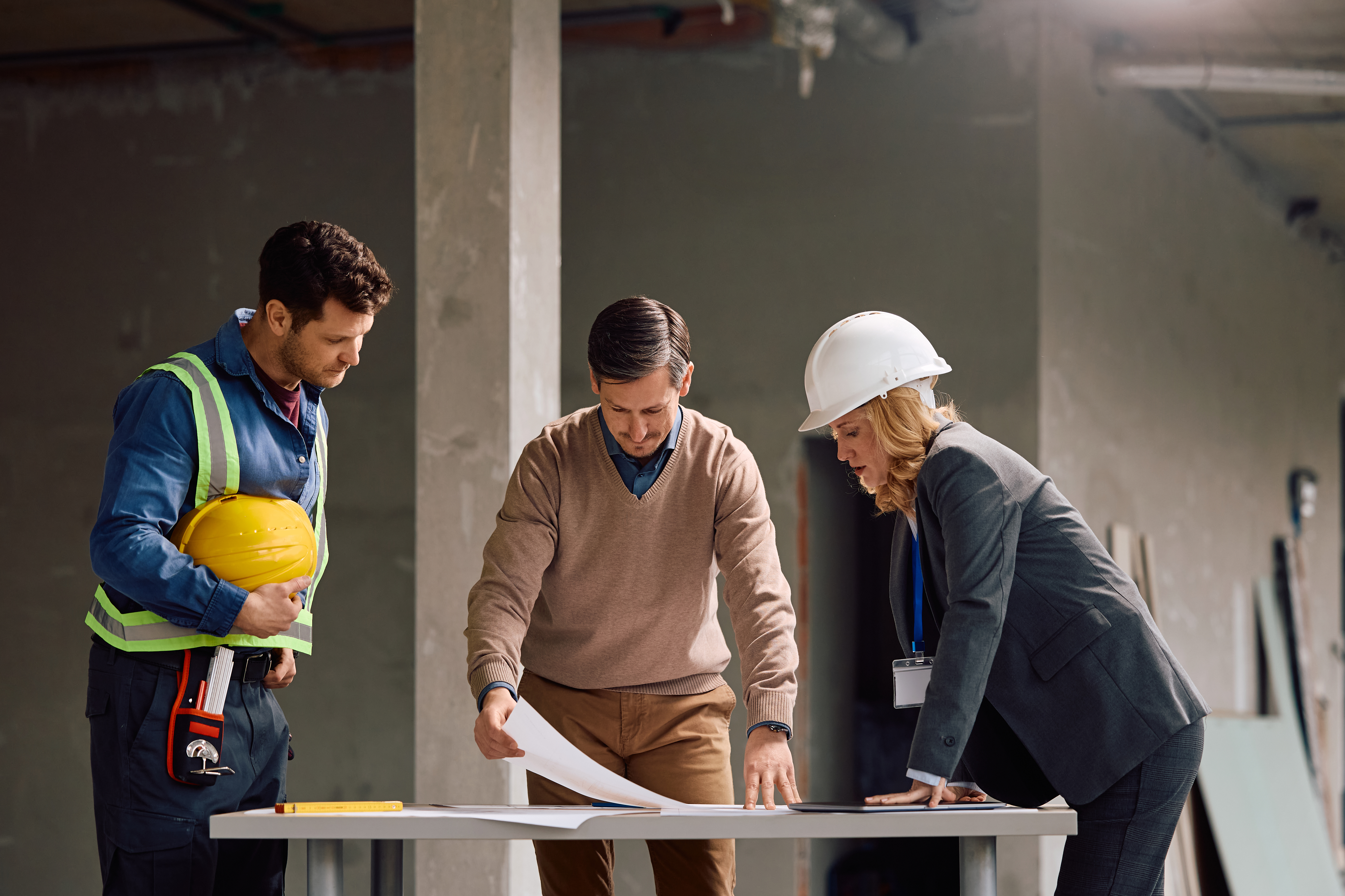 Building contractor cooperating with a worker and project manager while analyzing construction plans at building site.